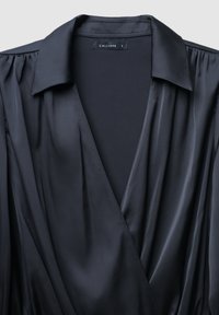 Schwarze Wickelbluse mit kragenartigem Ausschnitt, aus glattem, glänzendem Stoff und sanften Falten an den Schultern. Markenetikett sichtbar im Inneren.