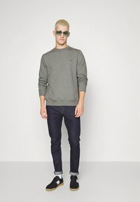 Grå genser med rund hals, mørke jeans og svarte joggesko. Tilbehør inkluderer grønne solbriller; modellen står mot en ensfarget bakgrunn.