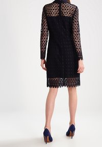 Schwarzes Spitzenkleid mit langen Ärmeln, taillierter Silhouette, floralem Muster und gewelltem Saum. Mit einem unsichtbaren Reißverschluss am Rücken abgeschlossen.