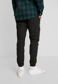 Zwarte cargobroek met elastische boorden, voorzien van zijzakken en een gladde textuur. Gecombineerd met witte sneakers en een groen geruit overhemd.