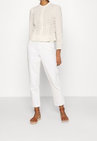 Weißes, tailliertes Strickjäckchen mit Lochmuster, langen Ärmeln und Rundhalsausschnitt, kombiniert mit weißen Cropped-Hosen und braunen Espadrilles.