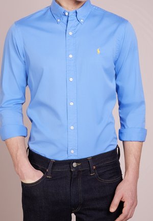 Homme portant une chemise bleu clair à manches longues avec poignets retroussés et un jean en denim foncé, une main dans la poche.