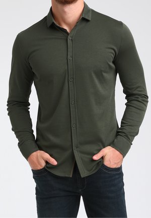 Mann trägt ein olivgrünes langärmliges Hemd mit Knöpfen und dunkle Jeans, steht mit den Händen in den Taschen vor einem schlichten hellen Hintergrund.