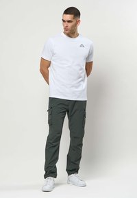 Mand står med hænderne bag ryggen iført hvid T-shirt med korte ærmer, mørke cargobukser med lynlåslommer og hvide sneakers på hvid baggrund.