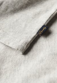 Jasnoszary materiał z przeszytym obszyciem i małą czarną metką z wyszytym złotym napisem "Superdry".