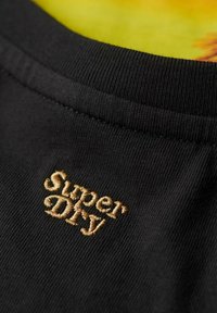 Cuello de camiseta negra con textura acanalada; presenta el nombre de la marca "Super Dry" bordado en dorado en la tela.