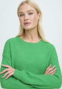 Pulover verde, moale, din material pufos, cu mâneci lungi și decolteu rotund. Are o croială lejeră și manșete cu dantelă.