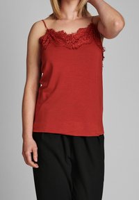 Top rosso senza maniche con pizzo sul colletto, spalline sottili e texture morbida. Indossato con pantaloni neri larghi.