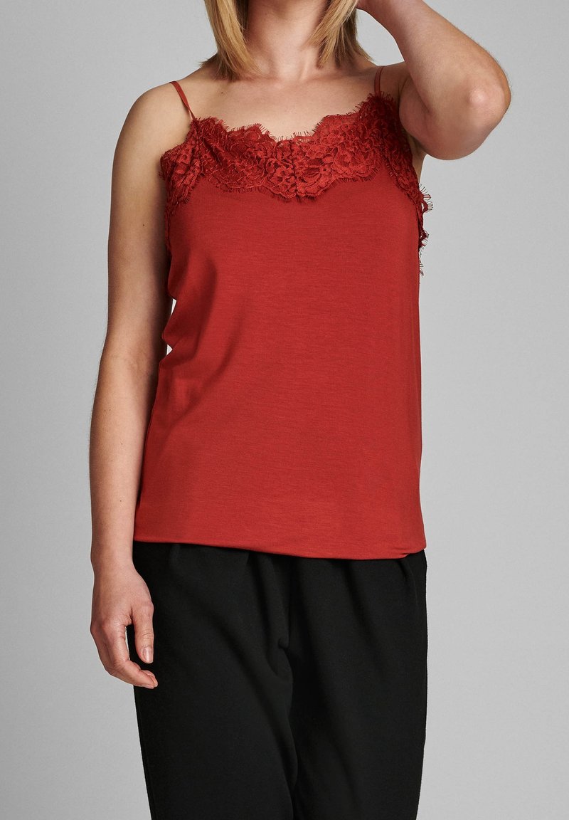 Top rosso senza maniche con pizzo sul colletto, spalline sottili e texture morbida. Indossato con pantaloni neri larghi.