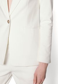 Biały żakiet o dopasowanym kroju, zapinany na jeden złoty guzik, z wąskimi klapami. Gładki materiał o subtelnej fakturze; zawiera boczne kieszenie.