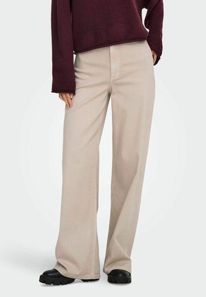 Person iført beige bukser med vide ben, sorte chunky sko og en langærmet mørkerød top, stående mod en lys baggrund.