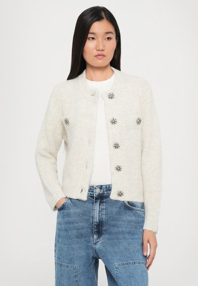 Kermankleurige gebreide cardigan met een ronde hals, acht decoratieve knopen en twee voorzakken. Draagt over een wit shirt en blauwe spijkerbroek.