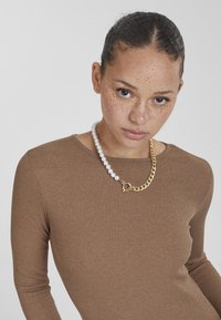 Maglia a maniche lunghe marrone a coste indossata con una collana che presenta perle bianche e una catena dorata, evidenziando varie texture e accenti metallici.
