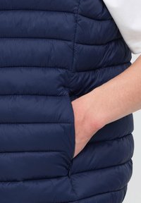 Mână în buzunarul lateral al unei veste matlasate bleumarin purtată peste o cămașă albă cu mânecă scurtă.