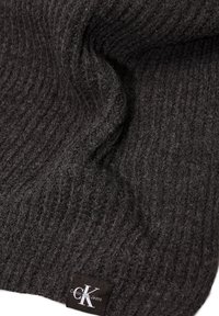 Sciarpa a coste di colore grigio scuro con una texture morbida. Presenta un piccolo etichetta con il marchio in testo bianco. Il tessuto cade dolcemente in pieghe.