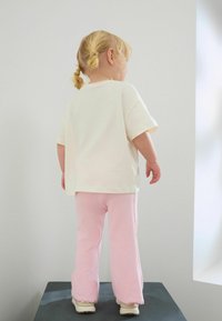 Jasno beżowa koszulka z krótkim rękawem i różowe prążkowane spodnie noszone przez dziecko, o luźnym kroju i casualowym designie, stojące na ciemnej powierzchni.