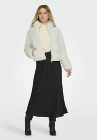 Giacca bianca gonfia con strisce verticali, texture a coste, collo alto e zip. Abbinata a un maglione crema e una gonna nera fluida.