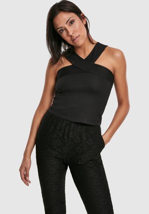 Vrouw draagt zwarte mouwloze top met kruisbandjes en zwarte kanten broek met hoge taille, poseert met één hand in de zak tegen een effen achtergrond.