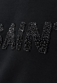 T-shirt in tessuto nero con lettere perlate che formano la parola "SAINT". Le perline sono lucide e di dimensioni variabili, creando una superficie testurizzata.