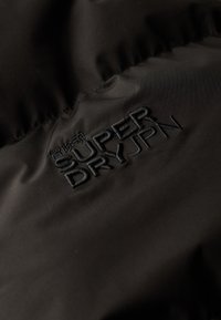 Fekete szövet, sima felülettel, a "SUPER DRY JPN" logó dombornyomott formában, kontrasztos színben. Az anyag könnyűnek és légáteresztőnek tűnik.