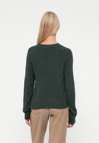 Tummanvihreä neulepaita, jossa on klassinen pyöreä kaula-aukko, ribbireunat ja pitkät hihat, valmistettu pehmeästä, teksturoidusta kankaasta. Yhdistetty vaaleanbeigeihin corduroy-housuihin.