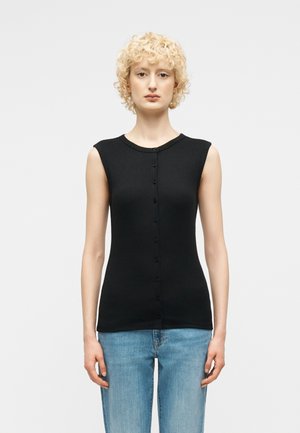 Blonde Person mit lockigem Haar, die ein ärmelloses schwarzes, geknöpftes Oberteil und hellblaue Jeans trägt, steht vor einem schlichten weißen Hintergrund.