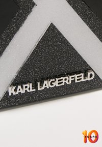 Materiale nero glitterato con un logo argentato "KARL LAGERFELD" e un design astratto bianco. Gli accenti includono una finitura strutturata e scritte in grassetto.