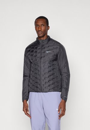 Muškarac nosi crnu quilted Nike jaknu sa zipom i svijetloružičaste Nike trenirke, stoji nasuprot obične bijele pozadine.