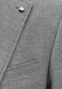 Gray textured blazer fabric featuring a close-up of a black button and a subtle pocket detail. The weave is small and tightly-knit.