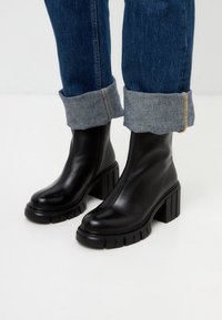 Zwarte leren enkellaarsjes met een chunky blokhak, voorzien van een zachte textuur en een minimalistisch ontwerp, gecombineerd met opgerolde denim jeans.