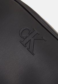 Černý kožený doplněk s texturovaným, vyraženým logem "CK". Hladký, matný povrch s jemným zrnitým vzorem, navržený s zaoblenými hranami.