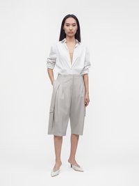 Jeune femme debout portant une chemise blanche boutonnée avec les manches retroussées, un pantalon beige taille haute et court, et des talons blancs pointus.