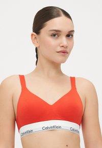 Červená bavlněná braletka s designem do V, širokými ramínky a bílým páskem s černým potiskem "CalvinKlein". Hladká textura a přiléhavý střih.