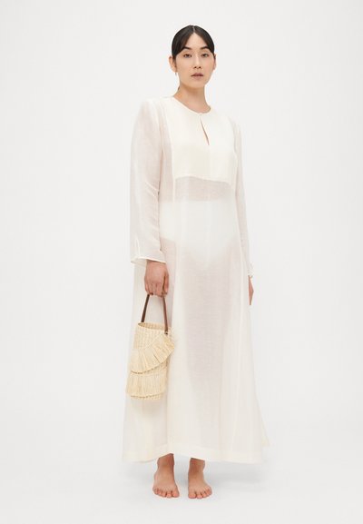 Μακρύ, διάφανο λευκό φόρεμα με άνοιγμα στο λαιμό και μακριά μανίκια. Συνοδεύεται από πλεκτή τσάντα άχυρου με διακοσμητικά κρόσσια.