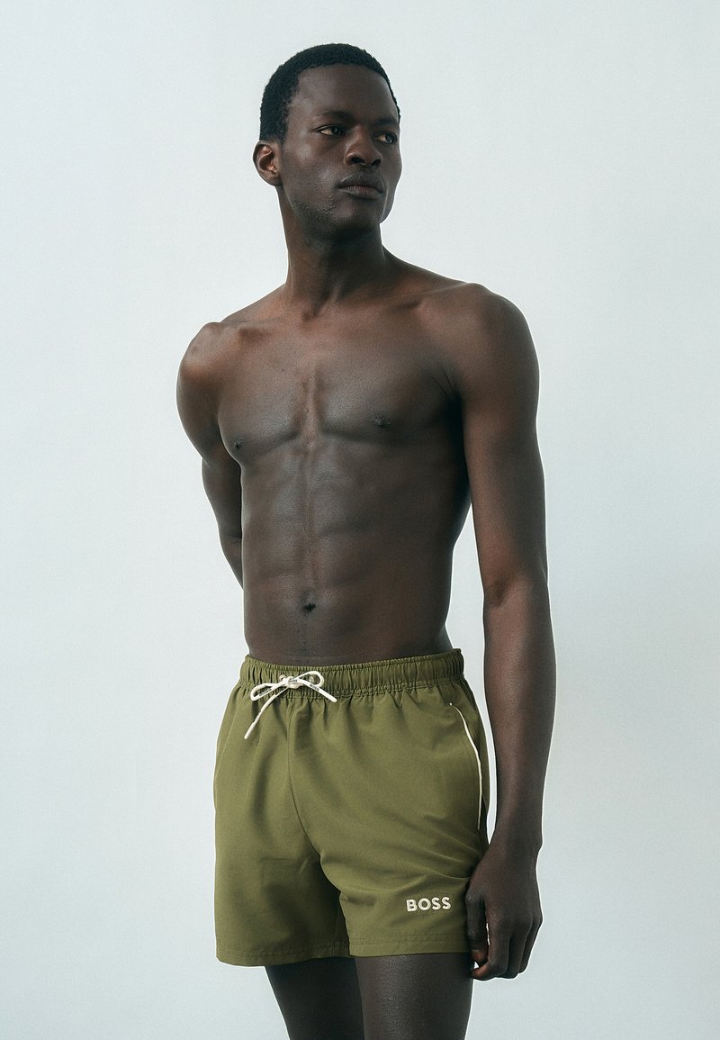 Άνδρας με σκούρο δέρμα χωρίς πουκάμισο, φοράει ελιά πράσινα μαγιό με το λογότυπο "BOSS", στέκεται σε ένα απλό φωτεινό φόντο.