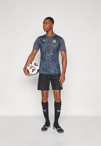 Jersey deportivo Puma en azul oscuro con patrones abstractos, logo dorado, combinado con pantalones cortos y calcetas negras, sosteniendo un balón de fútbol.