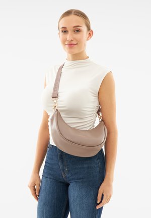 Braune Leder-Crossbody-Tasche mit runder Form, goldener Branding und verstellbarem Tragegurt. Kombiniert mit einem eng anliegenden, hellen Oberteil und Jeans.