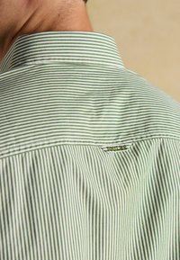 Zeleno-biele pruhované tričko s prehnutým golierom, ktoré má malú nálepku na zadnej strane krku a hladkú bavlnenú textúru.