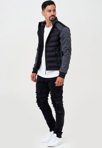 Svart dunstjakke med grå ermer, glidelås og ribbestrikkede mansjetter. Bæres over en hvit skjorte og kombinert med slitte svarte jeans og hvite sneakers.