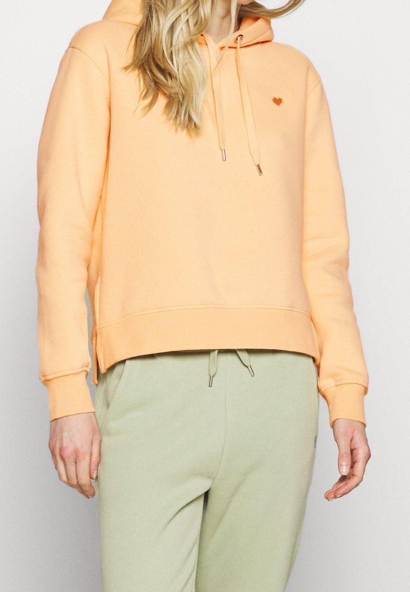 Hoodie broskvové barvy s malým srdíčkovým logem, s kapucí na šňůrku a bočními rozparky. Měkký bavlněný materiál, volný střih, kombinovaný se zelenými kalhotami.