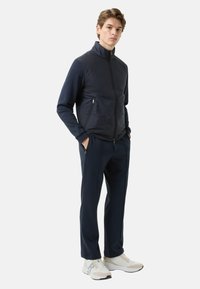 Navy zip-up jacket with a high collar, paired with matching navy trousers and light-colored sneakers. Textured fabric and subtle design details.
