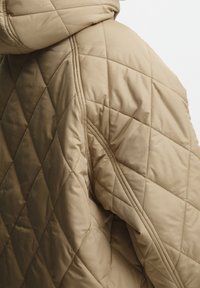 Beige kuddad jacka med huva, med diamantmönster och texturerad tyg. Axeln har kontrasterande sömnadsdetaljer.