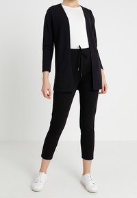 Persoon in een witte top, zwarte open voorkaart cardigan, zwarte drawstringbroek en witte sneakers, staand tegen een effen lichte achtergrond.