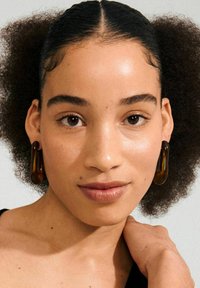 Orecchini a cerchio in resina marrone con una finitura lucida, caratterizzati da una forma curva e allungata. La modella ha capelli ricci e indossa un top nero.