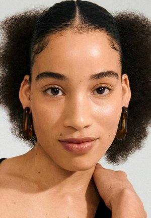 Bruine hars hoepel oorbellen met een glanzende afwerking, met een gebogen, langwerpige vorm. Model heeft krullend haar en draagt een zwarte top.