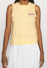 Gul ärmlös bomullstop med en rund Levi's-logotyp i rosa och svart framtill, med rund halsringning och en figurnära silhuett.