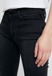 Vaqueros de denim negro con un diseño ajustado, que presentan un bolsillo frontal, costuras sutiles y un cierre de botón. Textura suave con minimalismo en los adornos.