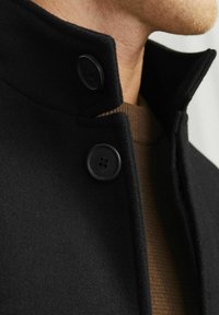 Abrigo de lana negro con un alto cuello, con dos botones negros. Debajo, se asoma un suéter marrón de canalé. Textura suave.