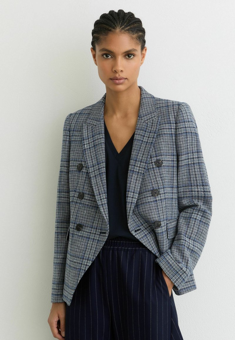 Vrouw met gevlochten haar, gekleed in een blauw geruit, double-breasted blazer over een zwarte V-hals shirt en marineblauwe broek met krijtstrepen.