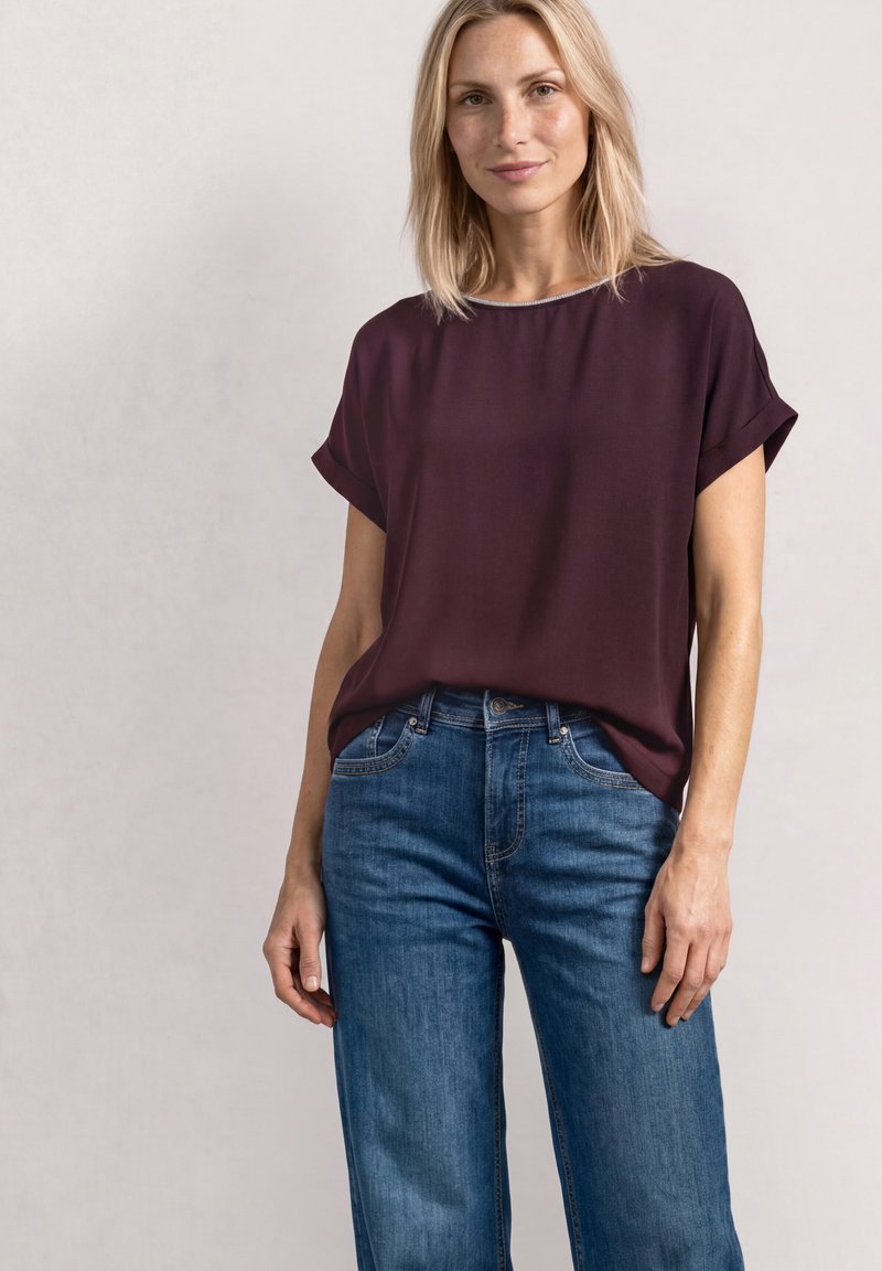 Femme blonde portant un chemisier ample à manches courtes violet foncé, rentré dans un jean bleu taille haute, debout devant un fond clair uni.